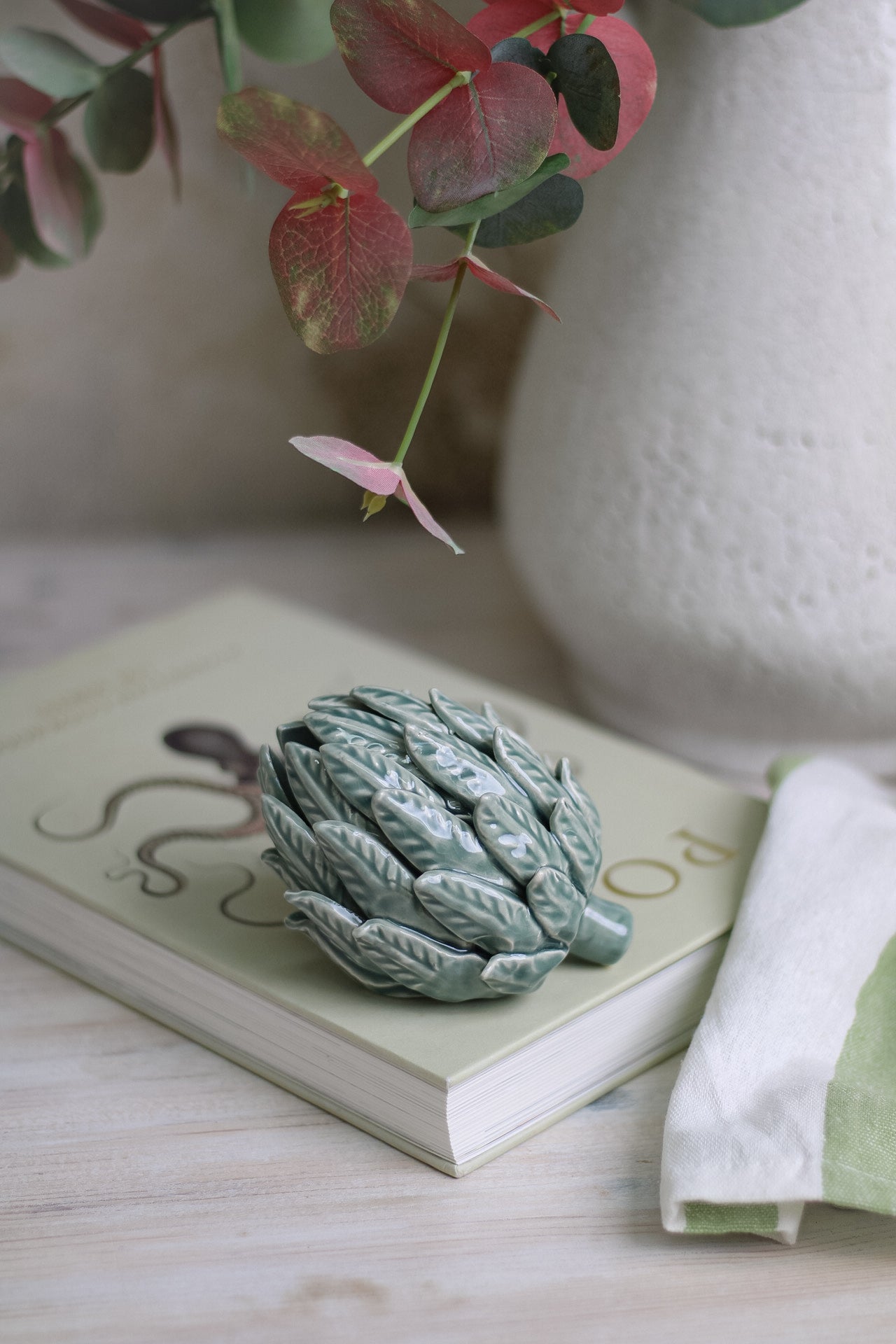 Mint Ceramic Artichoke resting on top of a coffee table book next to white vase