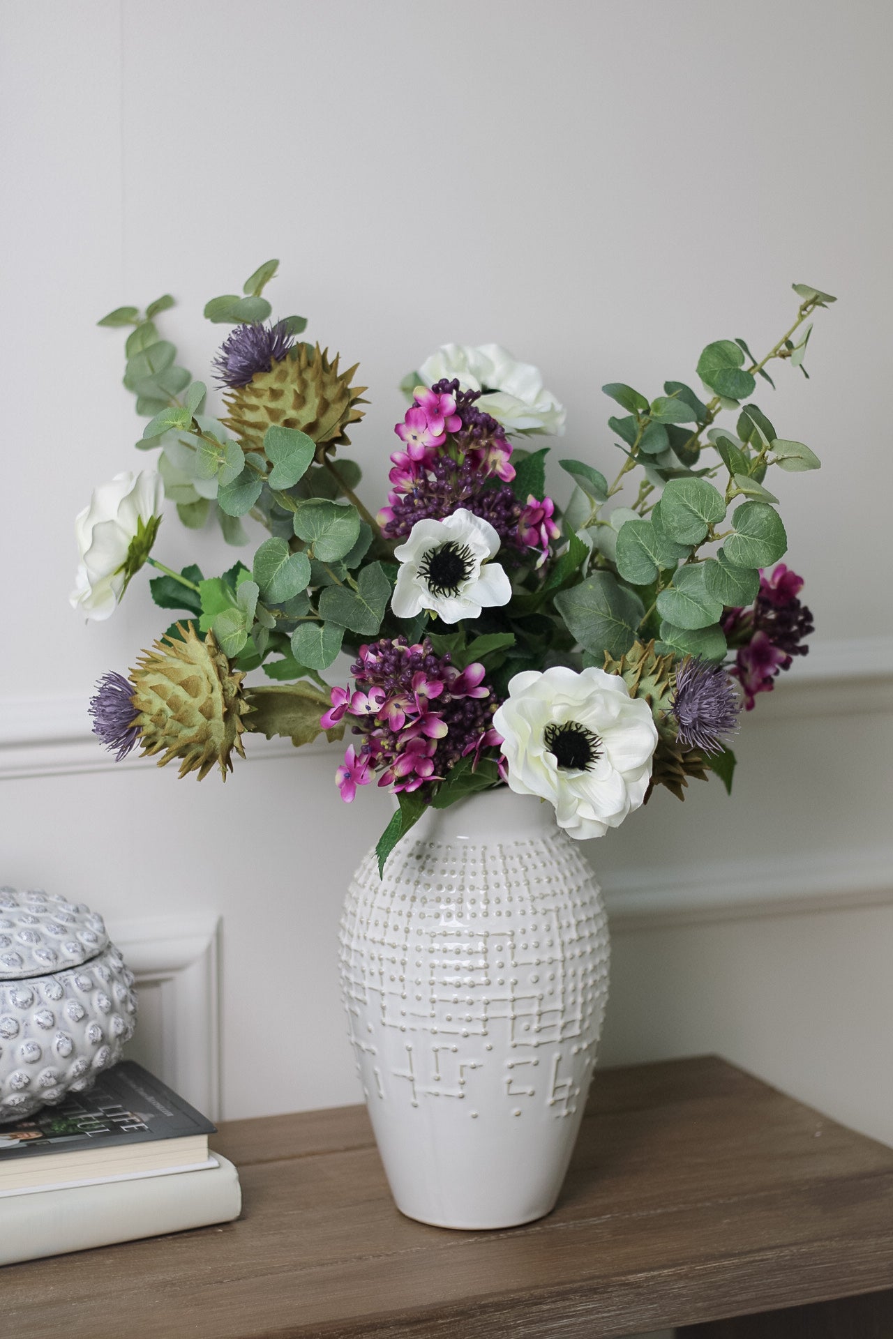 white ceramic vase with dotty pattern filled with faux eucalyptus, thistles and anemone stems.